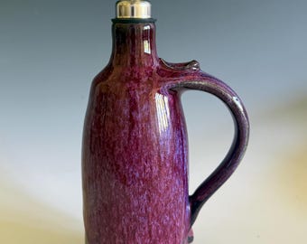May include: A ceramic oil cruet featuring a glossy, mottled purple glaze on the upper body and a solid dark black base. The vessel includes a curved handle on the side and a stainless steel pourer spout with a hinged metal cap at the top.