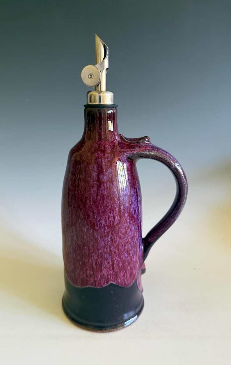 May include: A ceramic oil cruet featuring a glossy, mottled purple glaze on the upper body and a solid dark black base. The vessel includes a curved handle on the side and a stainless steel pourer spout with a hinged metal cap at the top.