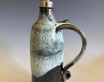 May include: A ceramic oil cruet featuring a two-tone glaze with a dark brown base and a speckled light blue top. The vessel includes a curved handle with a decorative scroll at the bottom and a stainless steel pour spout with a hinged lid.