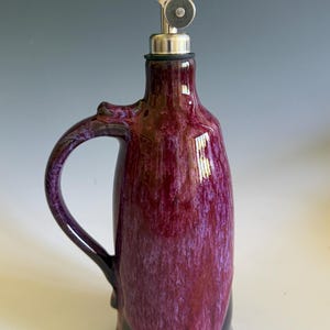 May include: A ceramic oil cruet featuring a glossy, mottled deep purple and magenta glaze with a smooth finish. The vessel has a curved handle on one side and is topped with a stainless steel pourer spout that includes a small circular metal cap.