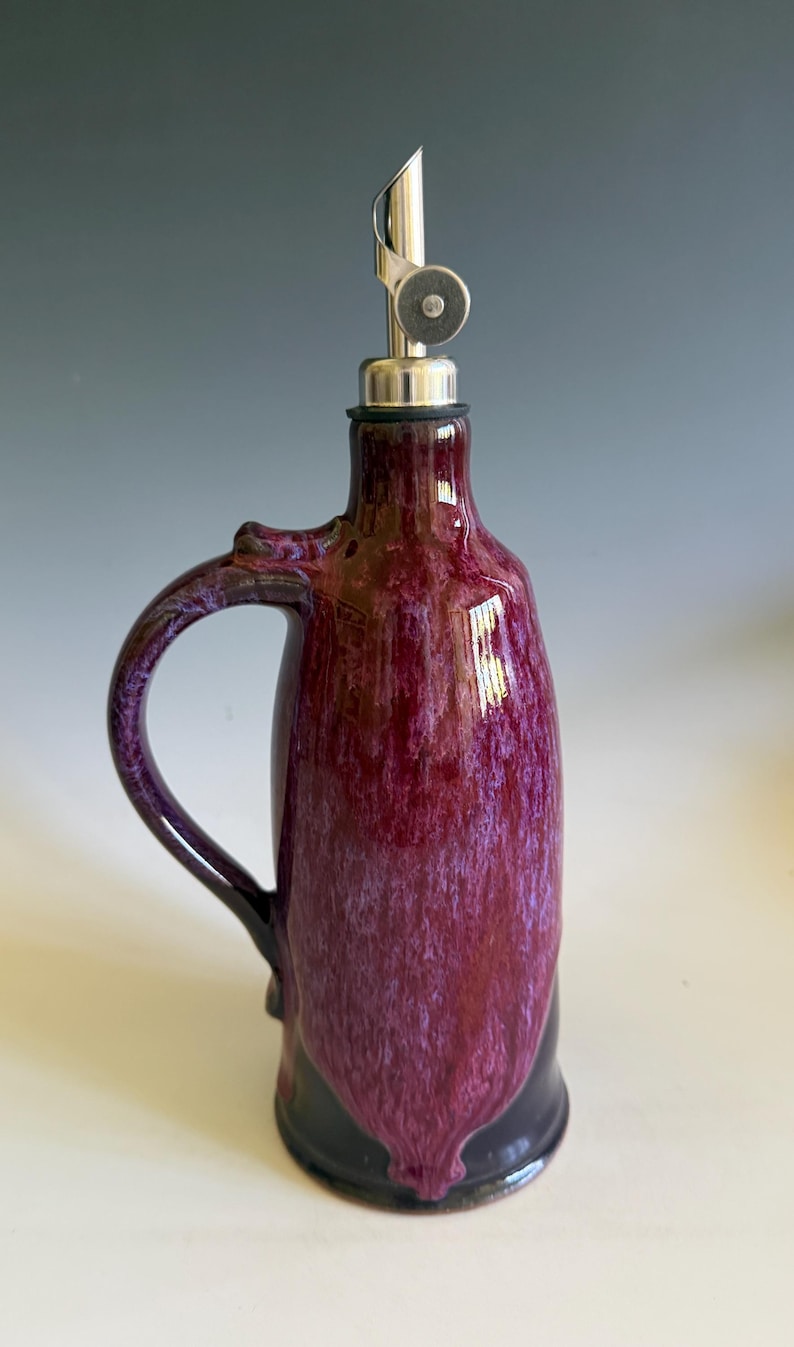 May include: A ceramic oil cruet featuring a glossy, mottled deep purple and magenta glaze with a smooth finish. The vessel has a curved handle on one side and is topped with a stainless steel pourer spout that includes a small circular metal cap.