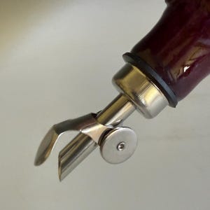 May include: A stainless steel bottle pourer with a hinged flap mechanism attached to a dark purple glass bottle neck. The metal spout features a circular side disc and a black rubber seal ring connecting the pourer to the bottle opening.