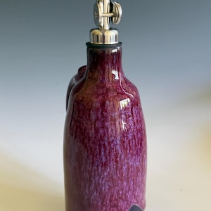 May include: A ceramic oil cruet featuring a glossy, mottled magenta and purple glaze that transitions into a solid dark base. The bottle is topped with a silver-colored metal pour spout with a hinged cap and a black rubber seal at the neck.