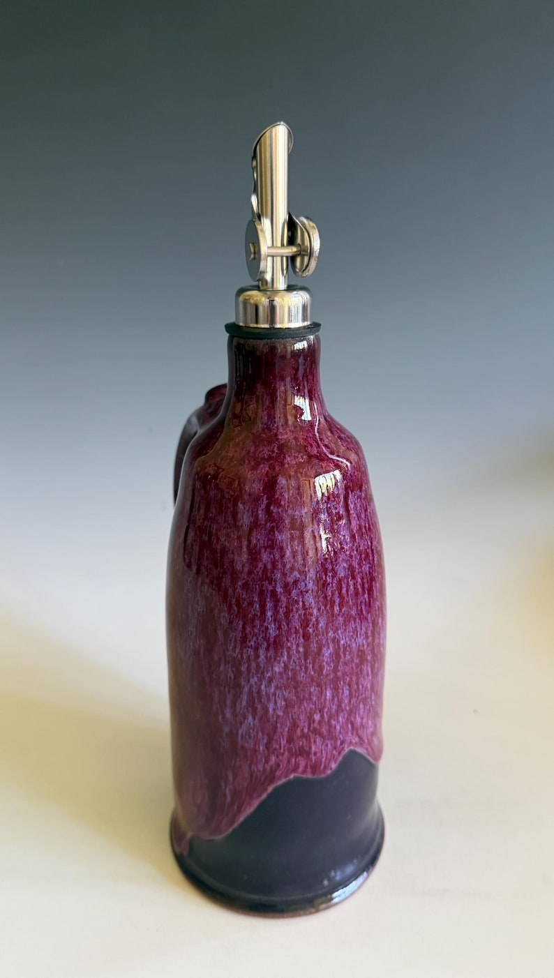 May include: A ceramic oil cruet featuring a glossy, mottled magenta and purple glaze that transitions into a solid dark base. The bottle is topped with a silver-colored metal pour spout with a hinged cap and a black rubber seal at the neck.
