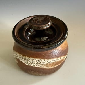 May include: A small, round ceramic jar with a lid. The jar is primarily brown with a white, crackled band around the middle. The lid and top rim are a darker brown. The jar has a small, round knob on top of the lid.