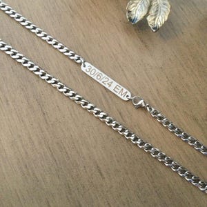 Peut inclure: Un collier chaîne argenté avec un design à maillons gourmette. Le collier comporte une étiquette rectangulaire avec le texte gravé "30/6/24 EM". La chaîne est présentée sur une surface en bois.