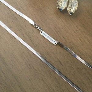 Puede incluir: Un collar de cadena plateada en espiga con un pequeño colgante rectangular. El colgante está grabado con las palabras "Always&Forever" y un símbolo de corazón. El collar se muestra sobre una superficie de madera.
