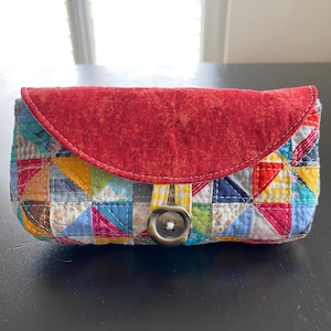 Op de afbeelding: Een rechthoekig gewatteerd tasje met een levendig patchworkpatroon van veelkleurige driehoeken en vierkanten. De flap is gemaakt van effen, gestructureerde rode stof en wordt gesloten met een grote, donkere metalen knoop met witte stiksels.