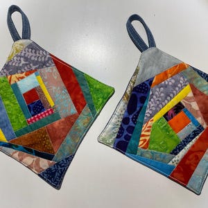 Op de afbeelding: Twee vierkante, gewatteerde pannenlappen met een patchwork-ontwerp. Elke pannenlap heeft een verscheidenheid aan kleurrijke stoffen stukken in tinten rood, groen, blauw en geel. Ze hebben denim lussen om op te hangen.