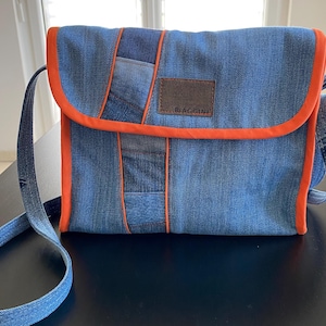 Könnte beinhalten: Eine blaue Jeans-Umhängetasche mit orangefarbenem Besatz und einem braunen Leder-Patch mit der Aufschrift "Giaggini".