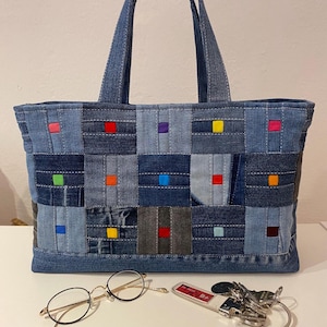 May include: A denim tote bag with a patchwork design. The bag is made from squares of various shades of blue denim, each accented with a small, colourful square. The bag has two handles and is on a white surface.