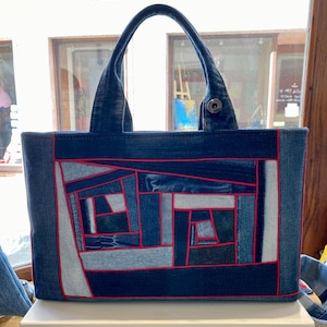 Può includere: Una borsa tote in denim con un design patchwork. La borsa ha una forma rettangolare con un motivo patchwork in tonalità denim blu, delineato con cuciture rosse. Ha due manici in denim e una chiusura con bottone.