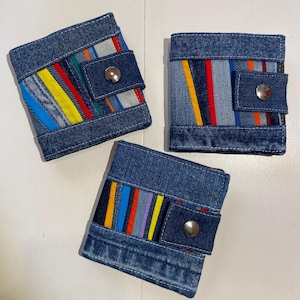 Peut inclure: Trois portefeuilles en denim avec des accents rayés colorés. Chaque portefeuille est doté d'un rabat avec une fermeture à bouton-pression argentée. Les portefeuilles sont fabriqués à partir de denim recyclé et de chutes de tissu.