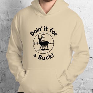 May include: A beige hooded sweatshirt with a front pocket. The sweatshirt features a graphic of a deer inside a crosshair, with the text "Doin' it for a Buck!" in a circular design. The hoodie is made of a soft material.