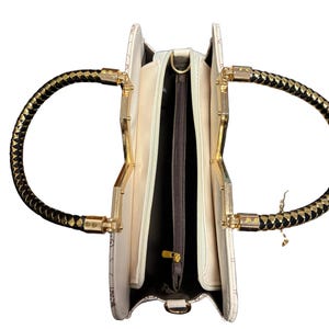 May include: Open ivory handbag with gold-toned hardware and black braided handles. The interior features multiple compartments with a zippered pocket. The bag has a decorative pattern and a gold zipper pull.