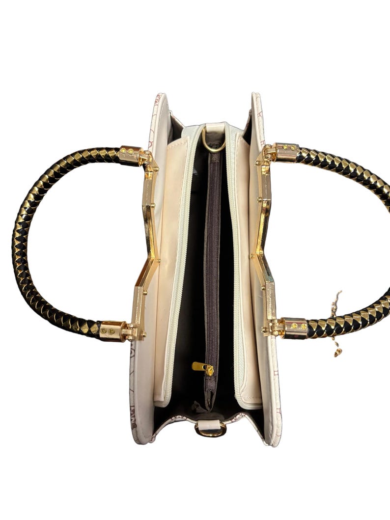 May include: Open ivory handbag with gold-toned hardware and black braided handles. The interior features multiple compartments with a zippered pocket. The bag has a decorative pattern and a gold zipper pull.