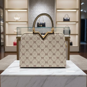 May include: A cream-colored handbag with a repeating brown pattern, gold-toned hardware, and a braided handle. The bag is displayed on a white marble surface in a well-lit boutique setting, showcasing other handbags in the background.