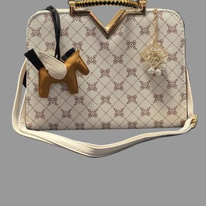 May include: Cream-colored handbag with a repeating pattern and gold accents. Features a gold handle, a decorative charm, and a brown pegasus-shaped charm. Includes a detachable white shoulder strap.