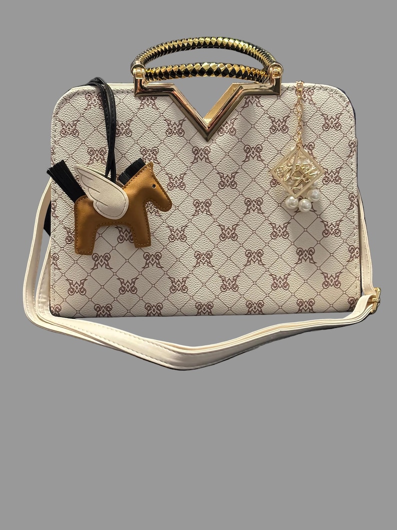 May include: Cream-colored handbag with a repeating pattern and gold accents. Features a gold handle, a decorative charm, and a brown pegasus-shaped charm. Includes a detachable white shoulder strap.