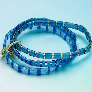 May include: A multi-strand bracelet composed of translucent blue and light blue rectangular glass beads interspersed with small gold-toned metallic spacers. A gold-toned circular ring clasp features a small cluster of tiny red stones.
