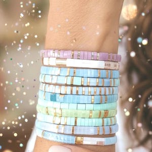 May include: A collection of colourful beaded bracelets in shades of blue, green, and lavender, with gold accents. The bracelets are stacked on a wrist, creating a layered look. The beads are small and rectangular, giving the bracelets a modern feel.