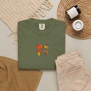 Puede incluir: Una camiseta de color verde oliva con un pequeño diseño bordado en el centro. El bordado muestra cuatro iconos de temática otoñal: una calabaza naranja, un farol amarillo, una seta roja con puntos blancos y una hoja de arce marrón.