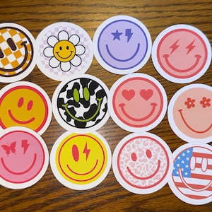 Puede incluir: Doce pegatinas redondas con diferentes caras sonrientes. Las pegatinas presentan diferentes diseños, incluyendo patrones de cuadros, florales, de vaca, corazones, estrellas, rayos y banderas americanas.