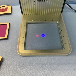 May include: A close-up of a silver machine with a square opening. Inside the opening, there are two small lights, one red and one blue.