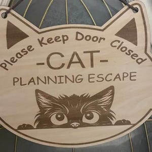 Pode incluir: Placa de madeira em forma de gato com o texto "Please Keep Door Closed - CAT - PLANNING ESCAPE". A placa apresenta um design de rosto de gato com uma ilustração de gato espreitando na parte inferior. Possui uma corda marrom para pendurar.