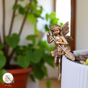 Può includere: Statuina di fata color bronzo con ali dettagliate e una piccola pala. La fata è appoggiata sul bordo di una fioriera bianca, con una pianta verde e frondosa sullo sfondo. La statuina ha un design delicato ed è un ornamento decorativo da giardino.