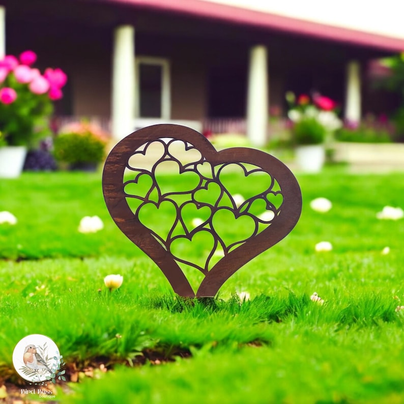 May include: A dark brown, heart-shaped garden stake with a cut-out design of smaller hearts. The stake is placed in green grass, with a blurred background of a house and flowers. The stake is approximately 12 inches tall.