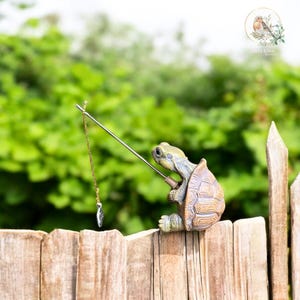 May include: A whimsical figurine of a turtle fishing, perched on a wooden fence. The turtle, with a detailed shell and green and brown coloring, holds a fishing rod with a silver charm. The background features lush green foliage.