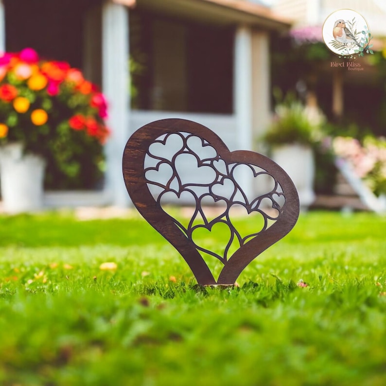 May include: A dark brown wooden heart-shaped garden stake with a cut-out design of multiple hearts. The stake is placed in green grass, with a blurred background of a house and flowers.