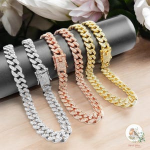 May include: Four iced-out cuban link chain necklaces in silver, rose gold, and gold. Each necklace features a secure clasp and is encrusted with sparkling stones. The necklaces are displayed on a black cylindrical stand.