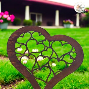 May include: A dark brown, heart-shaped garden stake with a cut-out design of smaller hearts. The stake is placed in a green lawn with a blurred background of a house and flowers. The stake is a decorative garden accent.