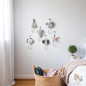 May include: A set of decorative wall hooks in a nautical theme. The hooks feature designs such as a sailboat, seashell, and starfish. Each hook has a silver metal hook. The hooks are mounted on a white wall in a bedroom.