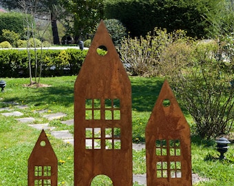 Könnte beinhalten: Drei rostige Metall-Gartenskulpturen in Form von Häusern. Jede hat ausgeschnittene Fenster und eine kreisförmige Öffnung in der Nähe der Oberseite. Sie stehen auf einem grasbewachsenen Rasen mit einem Hintergrund aus grünen Büschen und Bäumen.