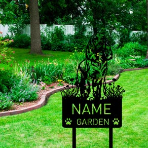 以下が含まれることがあります： 犬のシルエットと「NAME GARDEN」の文字が入った黒い金属製のガーデンサイン。サインは、背景に白いフェンスのある緑豊かな庭に設置されています。サインには足跡が含まれています。