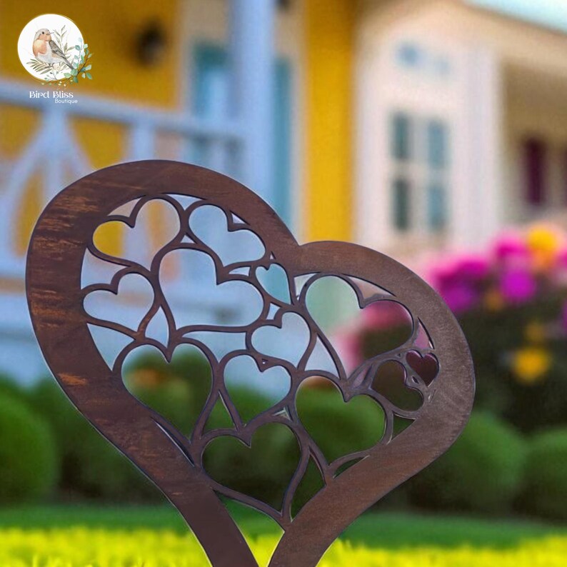 May include: A dark brown metal heart-shaped garden stake with a cut-out design of smaller hearts. The stake is placed in a grassy area, with a blurred background of a house and flowers. The logo "Bird Bliss" is in the upper left corner.