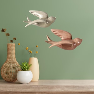 May include: Two ceramic bird wall decorations in flight. One is a muted grey, the other a warm brown. The birds have outstretched wings and are mounted on a green wall. Decorative vases with plants are in the foreground.