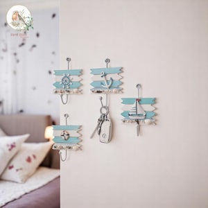 Può includere: Quattro attaccapanni a tema nautico, nei colori azzurro e bianco. Ogni gancio presenta un design diverso: un timone, un'ancora, un salvagente e una barca a vela. I ganci sono decorati con conchiglie e montati su una parete bianca.