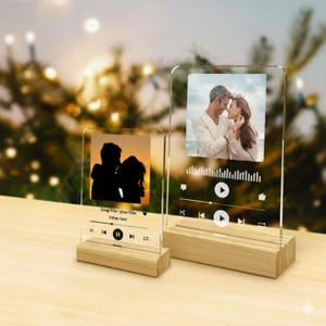 May include: Two clear acrylic photo frames with wooden bases. One frame shows a silhouette of a couple, the other a colour photo of a couple. Both frames have a music player interface design with play/pause buttons and song title text.