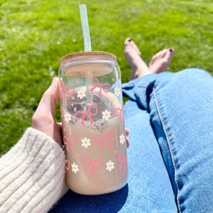 May include: A glass reusable cup with a straw, featuring a pink bow and white flower pattern. The cup is filled with a light brown beverage.
