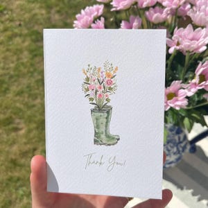 May include: A white card featuring a watercolour illustration of a green wellington boot overflowing with flowers in shades of pink, yellow, and purple. The words "Thank You!" are written in a script font. The card is held in a hand.