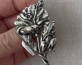 May include: A silver-toned brooch in the shape of a poppy flower with detailed leaves and a bud.