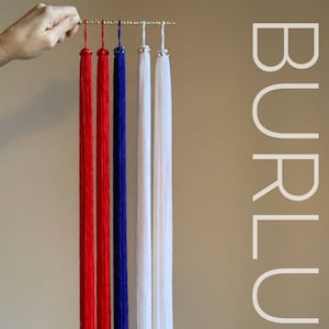 Puede incluir: Una mano sostiene una barra dorada con cinco largas borlas. Las borlas son rojas, rojas, azules y blancas. La palabra "BURLUXE" está impresa verticalmente en el lado derecho de la imagen.