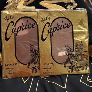 Puede incluir: Dos paquetes sellados de medias de nailon sin costuras de la marca "Caprice". El embalaje dorado presenta el nombre de la marca en una escritura estilizada, con las palabras "Seamless Nylons of Elegance" debajo. Los paquetes están sobre una superficie estampada.