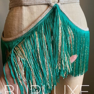 May include: A green and gold fringe trim with a gold ring detail. The fringe is attached to a tan fabric base.
