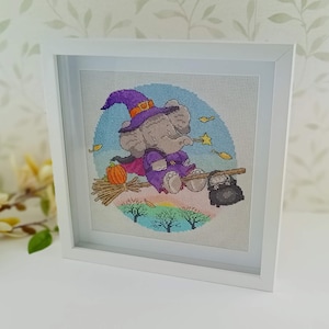 May include: A framed cross-stitch design featuring a purple and grey elephant dressed as a witch, flying on a broomstick with a cauldron. The elephant is wearing a purple hat and a purple dress. The background is a blue circle with a yellow star and orange leaves.