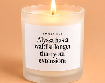 May include: A lit candle in a frosted glass jar with a white label featuring the black text: SMELLS LIKE Alyssa has a waitlist longer than your extensions. The candle emits a warm glow from the flame centered within the translucent glass container.